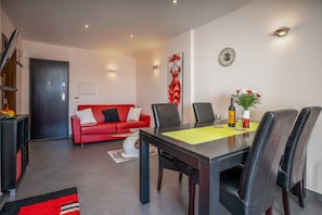 A dining area with a table, chairs, a red sofa, a clock, and a wall-mounted artwork.