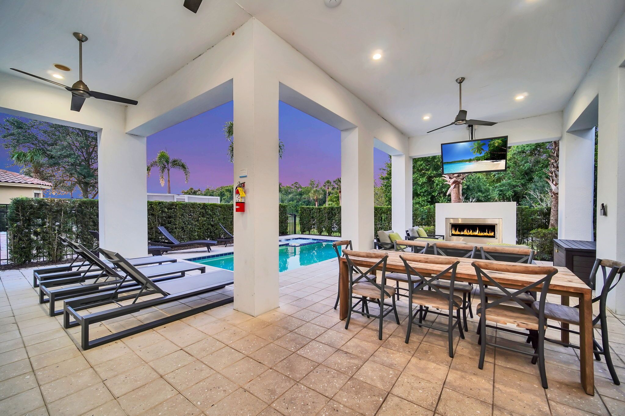 A poolside area with lounge chairs, a table with chairs, a flat-screen TV, and a fireplace.