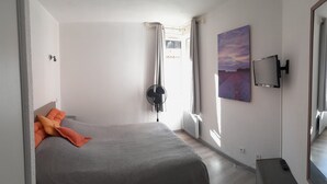 A bedroom with a bed, a standing fan, a TV mounted on the wall, and a window with curtains.