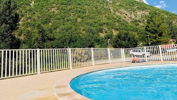 A swimming pool with a white fence, a clear blue sky, and a hill with greenery and rocky outcrops in the background.