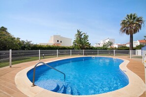 A swimming pool with a metal ladder, surrounded by a white fence, with buildings and a palm tree in the background.