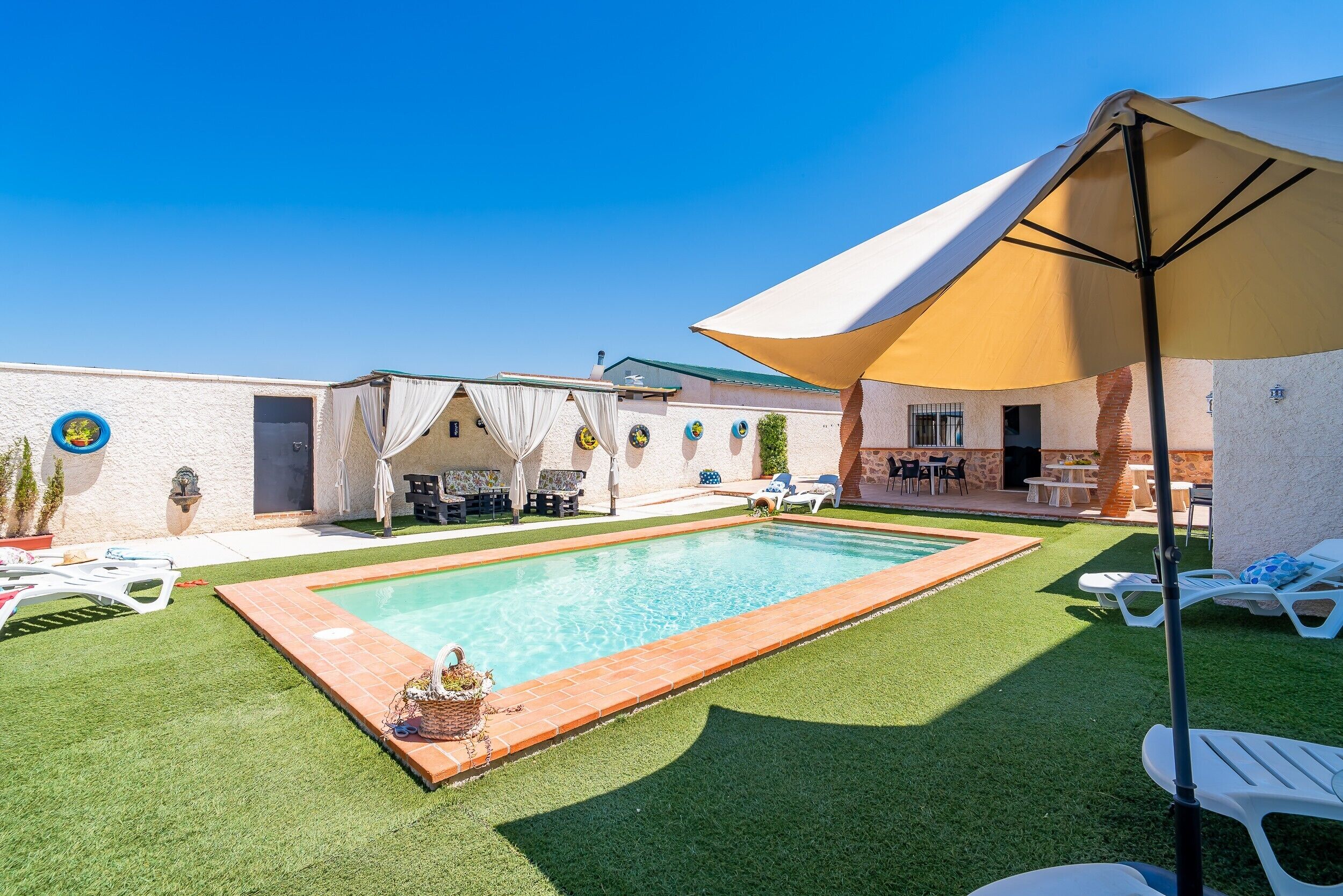 A swimming pool with a tiled border, surrounded by green artificial grass, and a covered patio area with outdoor furniture.