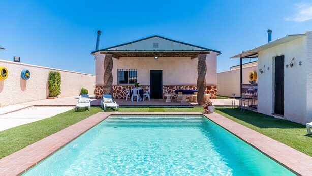 Una piscina con una zona prendisole, un'area salotto coperta e un piccolo edificio con una porta e finestre.