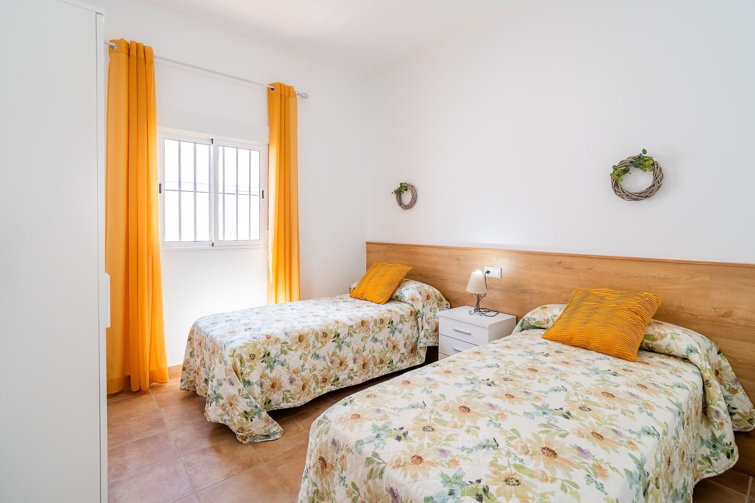 A bedroom with two beds, a window with curtains, a nightstand, and a wall with two wreaths.