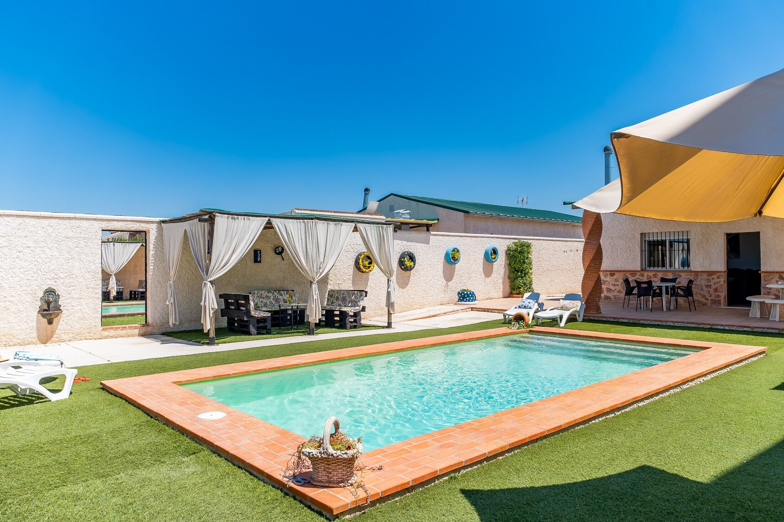 Ein Schwimmbecken mit Fliesenrand, umgeben von einer Rasenfläche und einer überdachten Terrasse mit Gartenmöbeln.