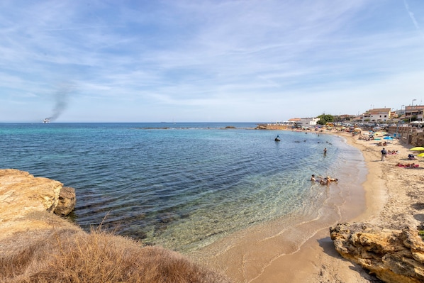 A beach with clear water, rocky shore, and buildings in the background.