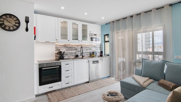 Una cocina moderna con armarios blancos, horno empotrado y un comedor con sofá y alfombra.