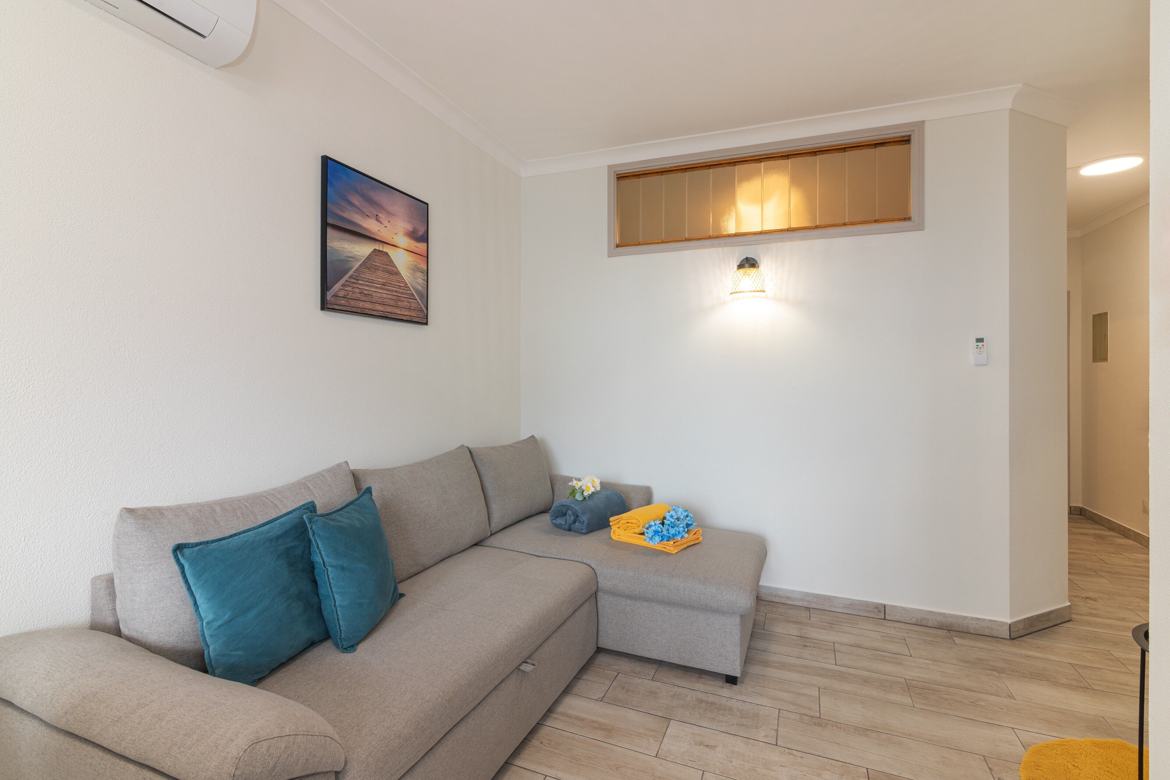 A living room with a beige sectional sofa, a painting, a wall-mounted light, and a wooden panel above a window.