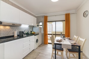 A modern kitchen with a washing machine, a dining area with a table set for two, and a view through a window with curtains.