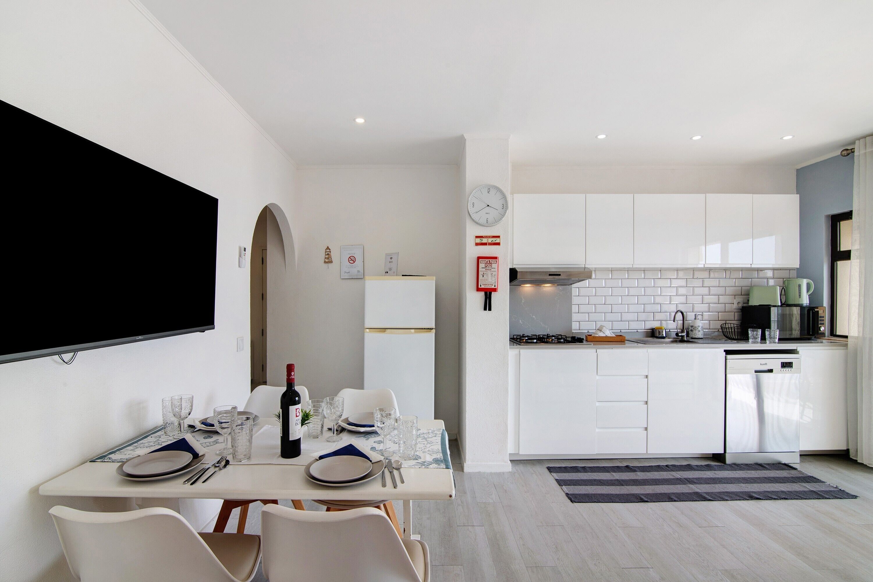 Una cocina moderna con armarios blancos, microondas y estufa. Hay un comedor con una mesa preparada para dos personas, incluyendo platos, vasos y cubiertos.
