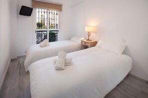 A hotel room with two beds, a TV mounted on the wall, a window with a view, and a bedside table with a lamp.