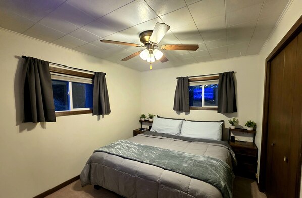 Un dormitorio con una cama, dos mesitas de noche, un ventilador de techo y dos ventanas con cortinas.