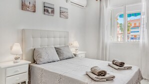 Une chambre à coucher comprenant un lit, deux tables de chevet, une commode, une fenêtre avec des rideaux et des œuvres d’art accrochées au mur.