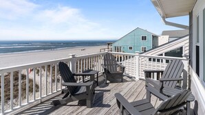Eine Holzterrasse mit Adirondack-Stühlen, Blick auf den Strand und ein hellblaues Gebäude.