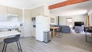 A modern kitchen with white cabinets, a microwave, a refrigerator, and a small trash bin. There is a dining area with a table and chairs, and a living area with a sofa and a TV. - Kings Little Secret four bedroom family home (Wānaka)