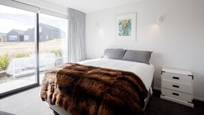 A bedroom with a large bed, a window with a view of houses, a framed picture on the wall, and a bedside table with drawers. - Modern family home with mountain views (Wānaka)