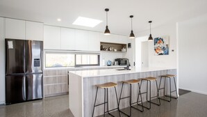 A modern kitchen with a stainless steel refrigerator, a white island with bar stools, pendant lights, and a large window providing natural light. - Modern family home with mountain views (Wānaka)