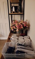 A room with a white door, a shelf with various items, a vase with pink flowers, and a container with blue objects.