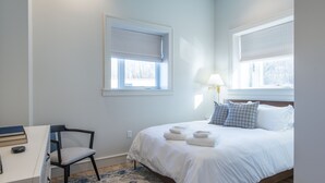A bedroom with a bed, two windows with blinds, a desk with a chair, and a lamp. - Barber Street Flats: #202 (Knoxville)