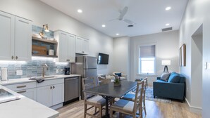 A modern kitchen with white cabinets, a dining table with chairs, a blue sofa, and a ceiling fan. - Barber Street Flats: #202 (Knoxville)