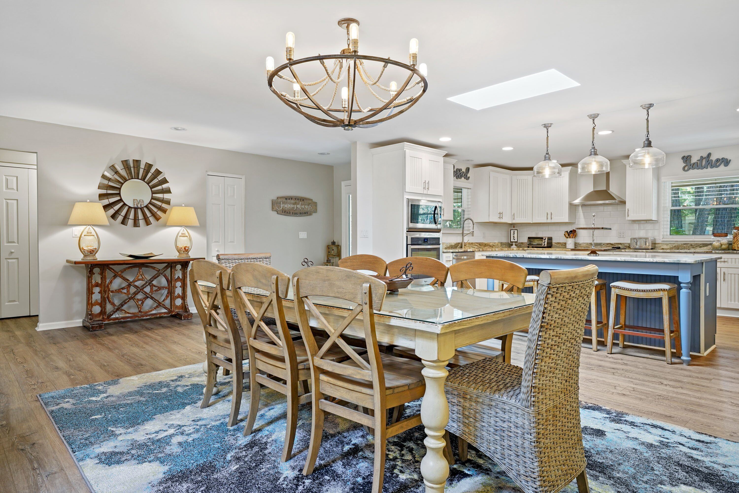 Una cocina amplia con armarios blancos, una isla central, iluminación colgante y un comedor con mesa de vidrio y sillas de madera.