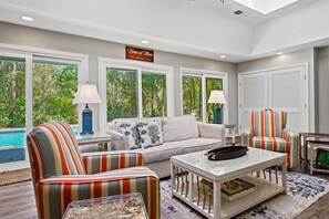 A spacious living room with a sofa, striped armchairs, a coffee table, and a view of a swimming pool through large windows.