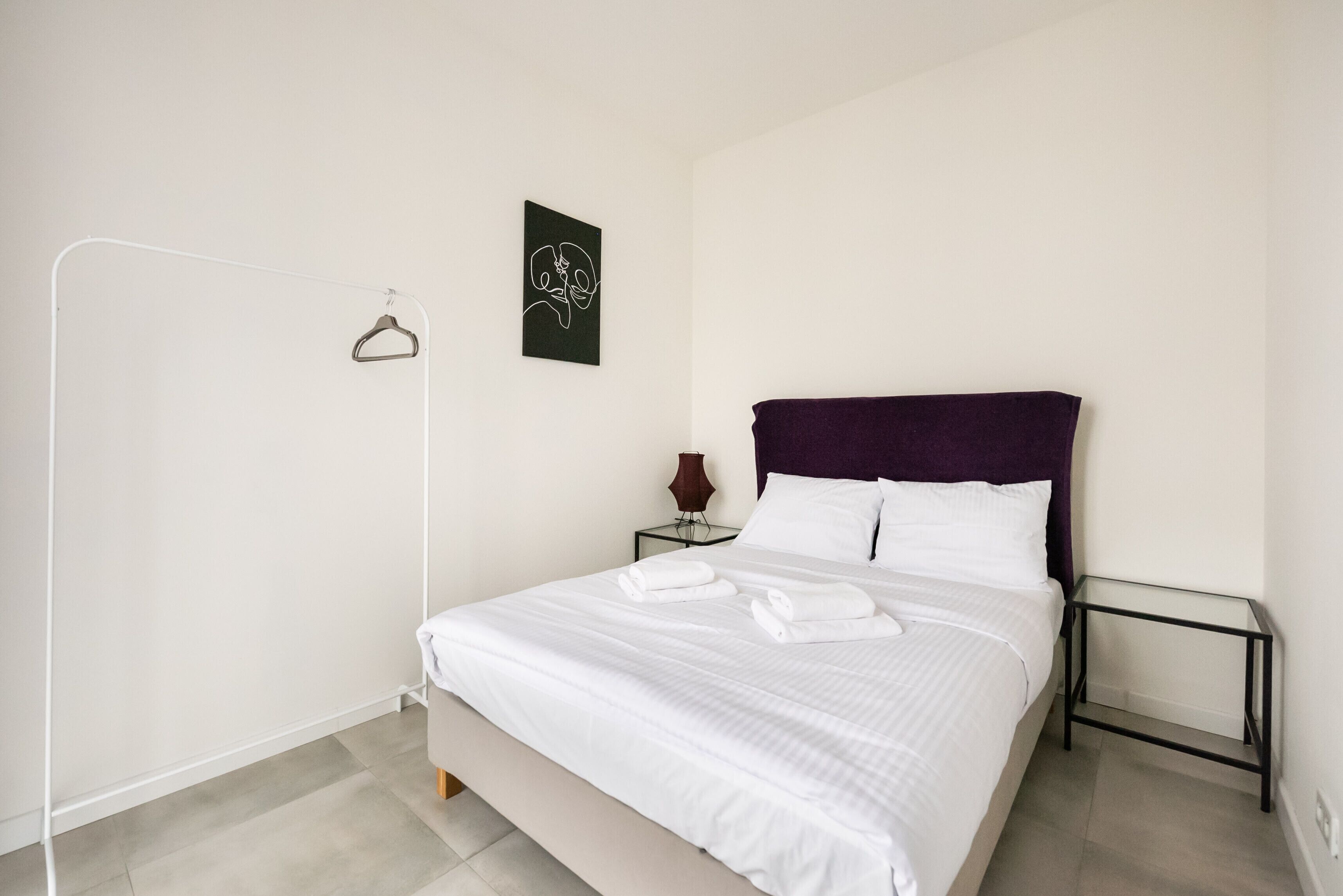 Una cama bien hecha con sábanas blancas y un cabecero morado, una mesita de noche con un jarrón, y una obra de arte colgada en la pared.