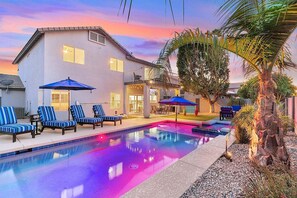 A pool area with lounge chairs and umbrellas, a large building with multiple windows, and a sunset sky. - Vacay In The Valley - Entertainers Backyard! (Gilbert)