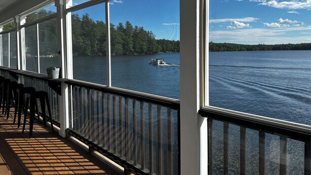 En inglasad veranda med stora fönster som vetter mot en sjö, en båt på vattnet och en skogbeklädd strand.