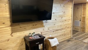 Una habitación con una televisión de pantalla plana grande montada en una pared de madera, una mesita con revistas y un control remoto, y un taburete de madera con una toalla blanca.