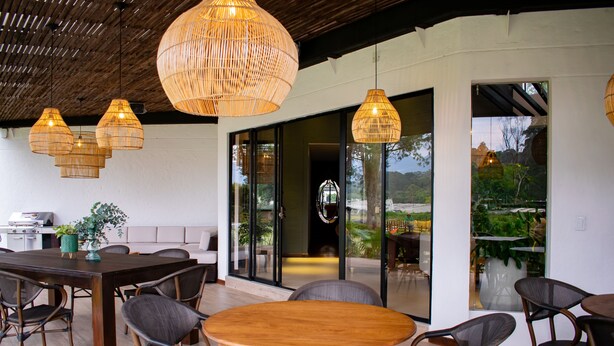 An outdoor dining area with wicker chairs and a wooden table, surrounded by large windows and a view of greenery. - Monarch Campestre (El Carmen de Viboral)