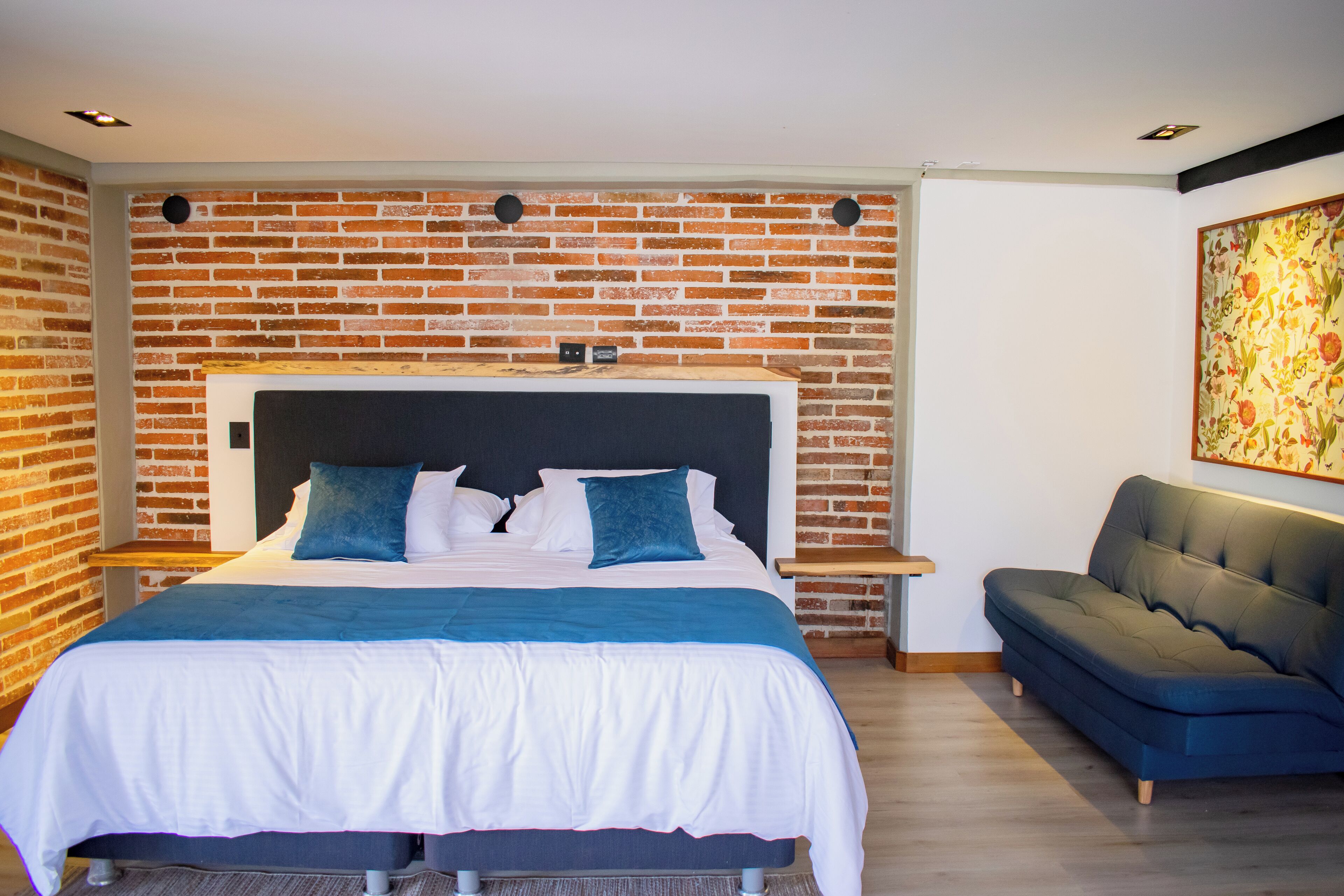 Une chambre d’hôtel moderne avec un mur orné d’un motif en briques, un grand lit doté d’une tête de lit en bois, un canapé bleu et une œuvre d’art accrochée au mur.