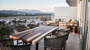 Un balcón con una mesa de madera y sillas de mimbre, con vistas a un paisaje urbano y montañas.