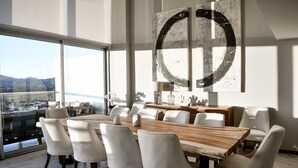 Un comedor moderno con una gran mesa de madera y sillas blancas tapizadas, un cuadro abstracto de gran tamaño en la pared, y vistas de la ciudad a través de amplios ventanales.