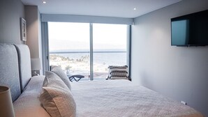 Un dormitorio con una cama grande, un televisor montado en la pared y vistas al océano a través de amplias ventanas.