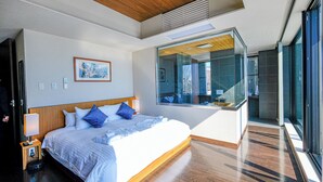 Ein modernes Hotelzimmer mit einem großen Bett, einer Duschkabine aus Glas und Blick auf den Poolbereich.