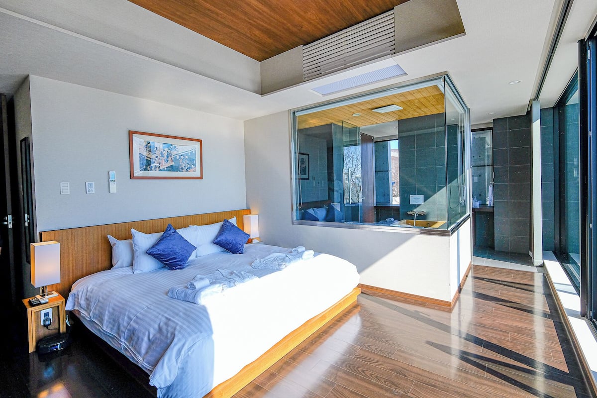 A modern hotel room with a large bed, a glass shower enclosure, and a view of the pool area.