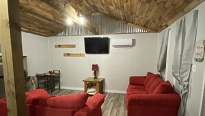 A room with a wooden ceiling, two red sofas, a small table with a lamp, a TV mounted on the wall, and an air conditioning unit. - James' Cabin (Gilbert)