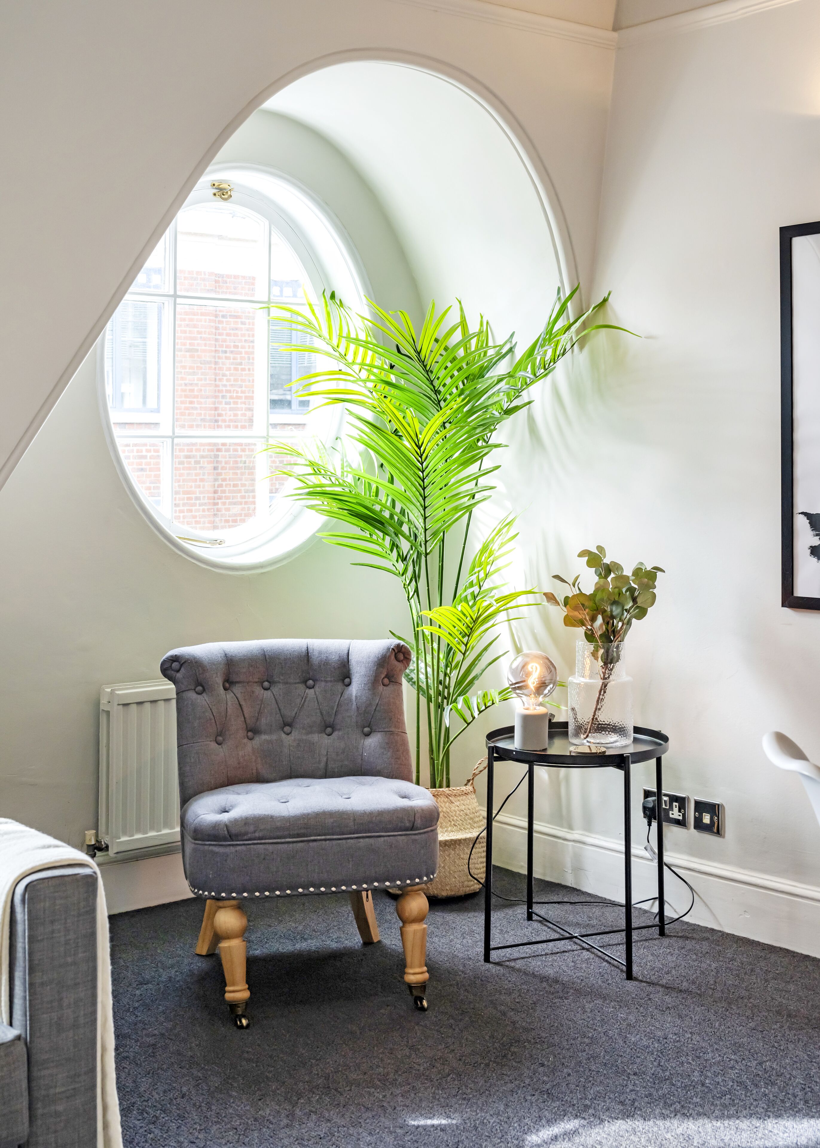 Une pièce comprenant un fauteuil à capitons, une grande plante en pot, une petite table avec un globe terrestre et des bougies, et une fenêtre donnant sur un bâtiment en briques.