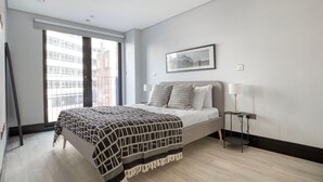 A modern bedroom with a large bed, a window with a view of buildings, a framed picture on the wall, and a bedside table with a lamp. - Exceptional Aldgate/Whitechapel 2BR w/ Garden, nr Station, by Blueground (London)