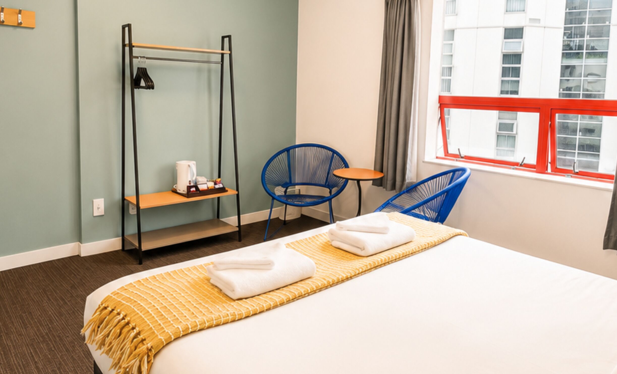A hotel room with a bed, a chair, a small table, a shelf, and a window with a view of buildings.