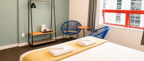A hotel room with a bed, a chair, a small table, a shelf, and a window with a view of buildings.