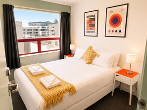Une chambre d’hôtel avec un lit, deux tables de chevet, une fenêtre avec des rideaux et deux tableaux encadrés accrochés au mur.