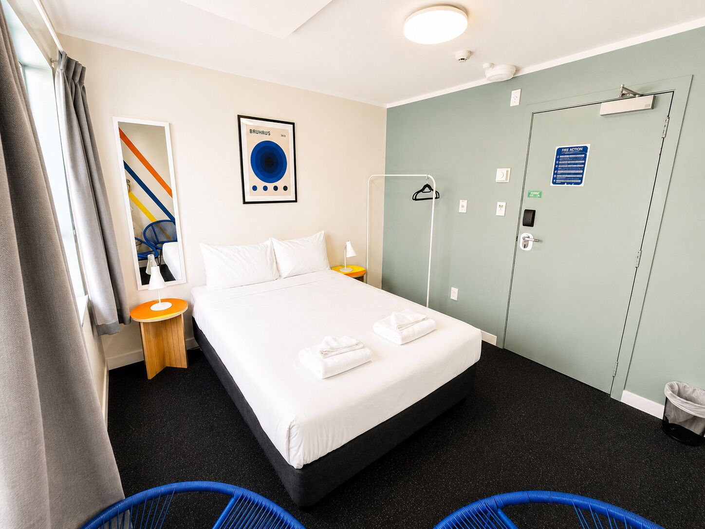 Habitación de hotel con cama, una silla, una mesita, una lámpara de pared, una puerta con un cartel y una ventana con cortinas.