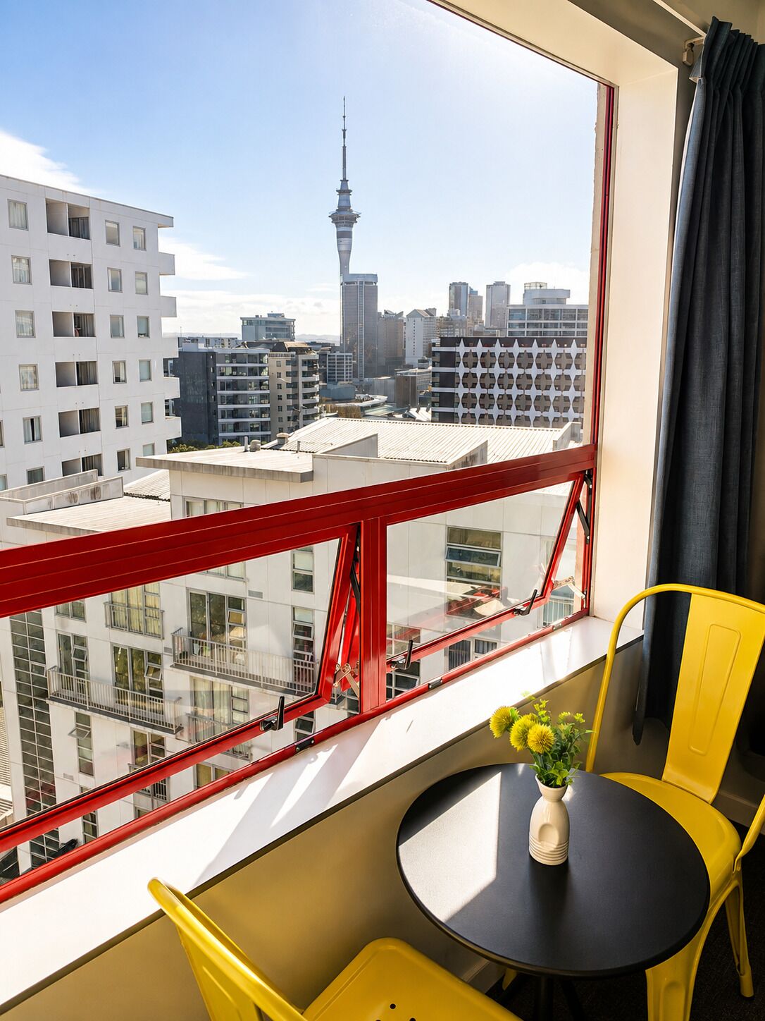 Una habitación con vista a un horizonte urbano, barandilla de balcón roja, una mesa redonda con un jarrón de flores y sillas amarillas.