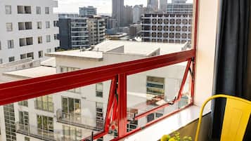 Una habitación con vista a un horizonte urbano, barandilla de balcón roja, una mesa redonda con un jarrón de flores y sillas amarillas.