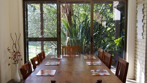 Una mesa de comedor con sillas, un florero con ramas y vista a árboles por una ventana.