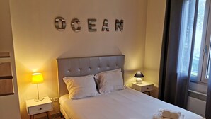 Siististi pedattu sänky, jossa on kaksi tyynyä, yöpöytälamppu ja seinälle kiinnitetty kyltti, jossa lukee ”OCEAN”.