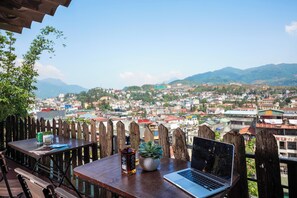Una mesa de madera con una laptop, una botella y una planta, con vistas a un paisaje urbano y montañas al fondo.