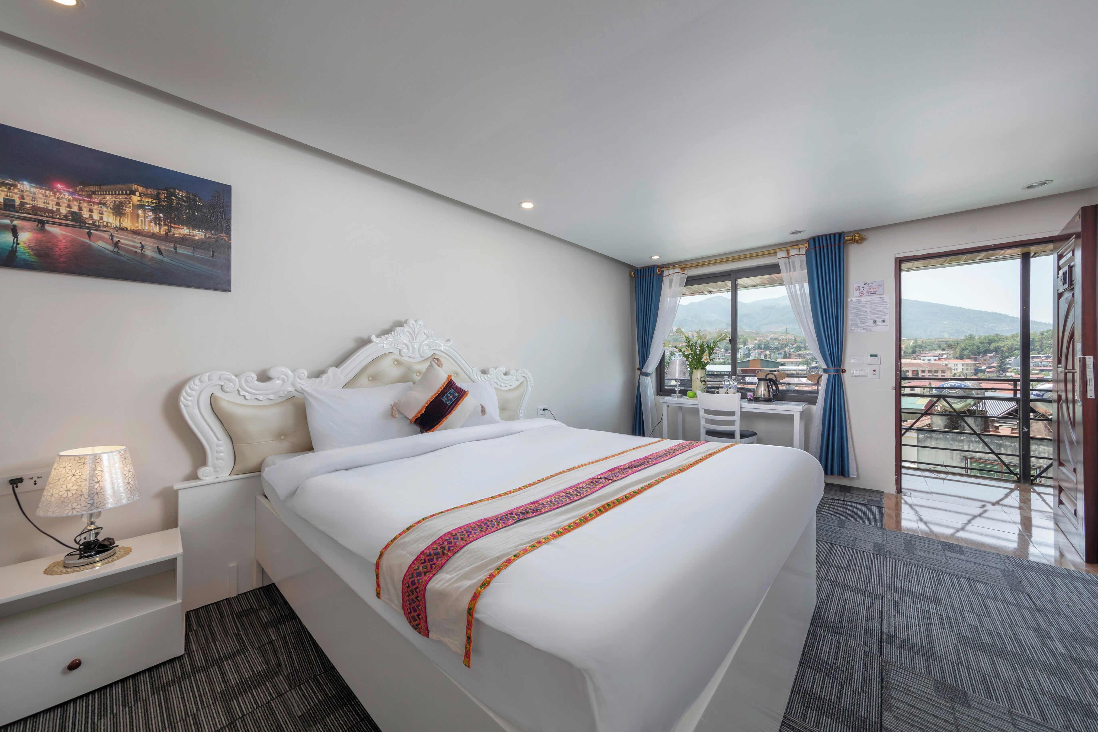 Una habitación de hotel con una cama grande, una mesita de noche, una lámpara, una ventana con cortinas y vistas a la ciudad.